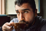 Fototapeta Miasto - Clear eyes handsome bearded man with brown hair with thoughtful look while holding a cup or mug and drinking tea or coffee in wooden studio or cofee shop. Close portrait