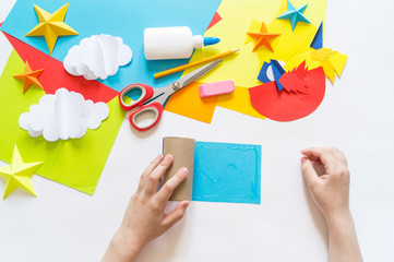 Wall Mural - hands of the child make a paper craft rocket. Cosmos clouds and stars colored paper. The creative process