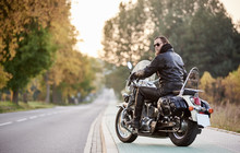 Back View Of Handsome Bearded Motorcyclist In Black Leather Jacket And Sunglasses Sitting On Cruiser Motorcycle On Blurred Background Of Straight Road Stretching To Horizon And Golden Autumn Trees.