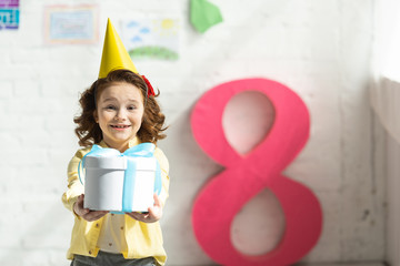 Wall Mural - adorable smiling kid in party cap holding present and posing near decorative pink number 8