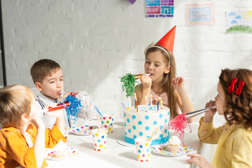 Wall Mural - kids sitting at table with cake and cheering with party horns during birthday celebration