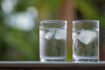 Transparent misted glasses with water and ice against a background of green bushes in different lighting