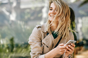 Portrait of happy young beautiful woman smiling and looking away, using mobile phone, sitting outdoor in the city at sunny day. Pretty female reading news on cellphone. Travel and technology concept