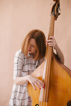 Beautiful Young Woman Musician Sitting On A Vintage Double Bass On A Beige Background In A Studio