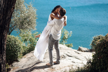 Wall Mural - Man holding woman in his arms wrapped around her waist and she is holding on to him. The wind is blowing her hair across her face, standing in a seascape near the mediterranea sea in Greece.