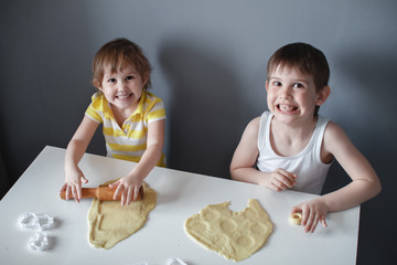 Wall Mural - Little cheerful children make shortbread. Rolls out the dough with a rolling pin on a white table. The preparation of the dessert