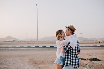 Young traveling guy in stylish hat and girl in trendy outfit cute kissing near the road at weekend. Adorable young woman gently embracing her handsome boyfriend and smiling on nature background