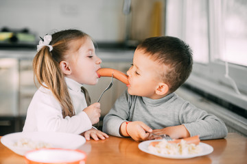 Wall Mural - children in the kitchen boy and girl eat sausages with pasta very fun and together share one