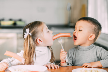 Wall Mural - children in the kitchen boy and girl eat sausages with pasta very fun and together share one