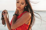 Fototapeta Na drzwi - close up of a beautiful asian filipino girl with smooth tan skin and her hair blowing in the wind while holding a sandal and wearing an orange sleeveless blouse on the sandy beach