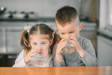 Children Boy And Girl In The Kitchen Drinking Water From Glasses