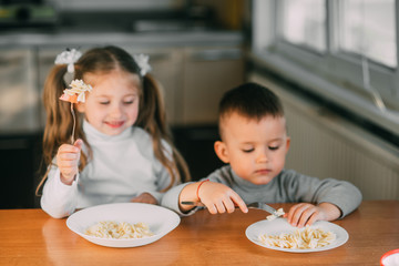 Wall Mural - Boy and girl children in the kitchen eating sausages with pasta is very fun and friendly