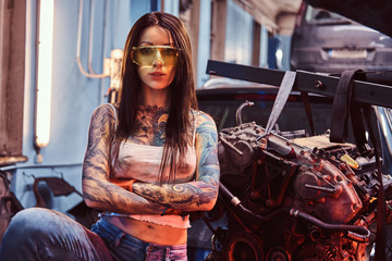 Female car mechanic holding a big hammer and posing next to a car engine suspended on a hydraulic hoist in the workshop.