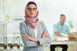 © Suzi Media  - Beautiful young working woman in hijab, suit and eyeglasses standing in office, smiling. Portrait of confident muslim businesswoman. Modern office with big window, man working at desk on background.
