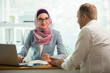 Canvas Print - Coworkers meeting in office. Stylish woman in hijab making conversation at desk with man in white modern office. Muslim businesswoman in eyeglasses interviewing man.