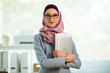 © Suzi Media  - Beautiful young working woman in hijab and eyeglasses smiling in office, holding folders and laptop in arms. Portrait of confident muslim businesswoman. Modern office with big window on background.