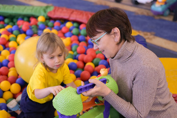 Grandson and grandmother playing together. Portrait of a blond boy in a yellow t-shirt. The child smiles and plays in the children's playroom.