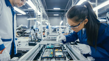 Shot Of An Electronics Factory Workers Assembling Circuit Boards By Hand While It Stands On The Assembly Line. High Tech Factory Facility.