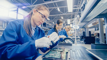 Female Electronics Factory Worker In Blue Work Coat And Protective Glasses Is Assembling Laptop's Motherboard With A Screwdriver. High Tech Factory Facility With Multiple Employees. 