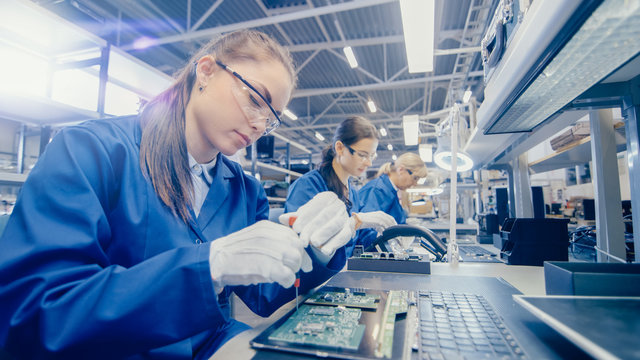 female electronics factory worker in blue work coat and protective glasses is assembling laptop's mo