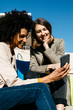 © Westend61 - Two happy women with shopping bags sitting outdoors using cell phone