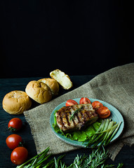 Grilled steak served on a plate, decorated with spices for meat, rosemary, greens and vegetables on a dark wooden background. Space for text.