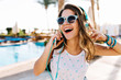 Close-up portrait of excited curly tanned girl in trendy sunglasses walking by swimming pool outside. Funny cheerful young woman in headphones enjoying music, while resting outdoor on summer resort.