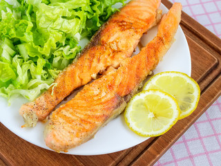 Fried salmon fillet on a white plate with fresh green lettuce and two slices of lemon. Healthy eating and low carb diet. Tasty seafood.