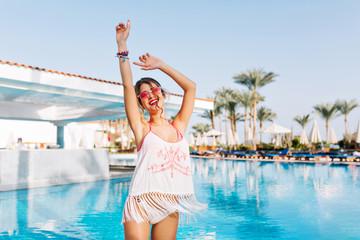 Beautiful slim girl in fringe shirt and white panties having fun near the warm blue water with palm trees on background. Cute young woman laughing and dancing at the pool on exotic resort in weekend