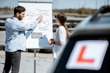 Male instructor showing traffic shemes to a young female student standing on the training ground at the driver's school