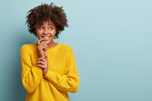 Optimistic Lovely Woman With Curly Hair, Keeps Hand Near Toothy Smile, Looks Aside, Has Dreamy Expression, Feels Shy, Wears Yellow Casual Jumper, Stands Against Blue Background. Copy Space Right