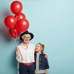 Vertical image of glad granny and small female child embrace, have nice relationships, celebrate holiday together, holds red balloons, enjoy birthday party, isolated on blue. Family portrait.