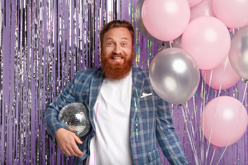 Wall Mural - Joyful handsome man throws party after promotion at work, holds disco ball and bunch of air balloons, smiles happily, wears elegant jacket, stands over purple tinsel curtain, enjoys loud music