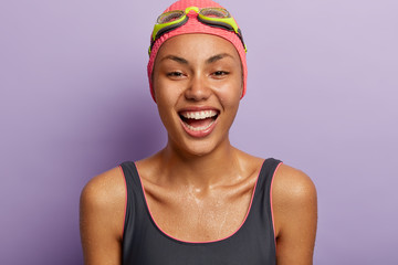 Wall Mural - Glad sportswoman professional swimmer wet after training in swimming pool, wears bathing suit, hat and goggles, shows white teeth, isolated over purple background. Summer time and active rest concept