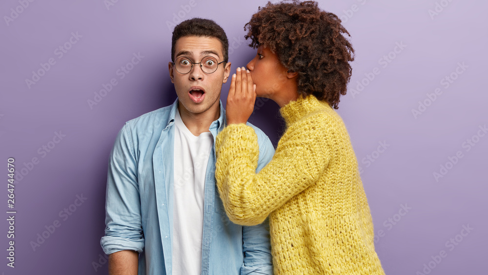 Photo of Afro American woman whispers secret to boyfriend, tells shocking news. Astonished young man keeps jaw dropped from surprisement, hears terrible rumors, isolated over purple background - obrazy, fototapety, plakaty 