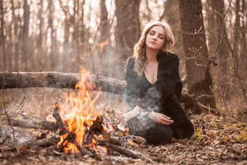 Wall Mural - Attractive young woman with black cape sitting in forest near bonfire. Eyes closed