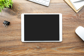 White tablet with black blank screen is on top of wood desk table with supplies. Top view, flat lay.