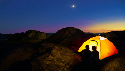 Wall Mural - young couple sitting near illuminated tent and looking at each other at beautirul evening in the mou