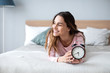 © BestForYou - Smiling young woman lying awake in bed with alarm clock, good morning.