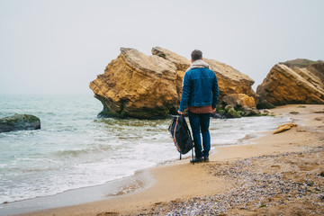 Wall Mural - traveler with a backpack standing near a rock against a beautiful sea with waves, a stylish hipster 