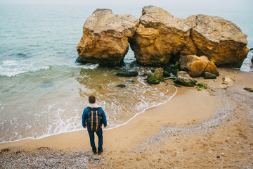 Wall Mural - traveler with a backpack standing near a rock against a beautiful sea with waves, a stylish hipster 