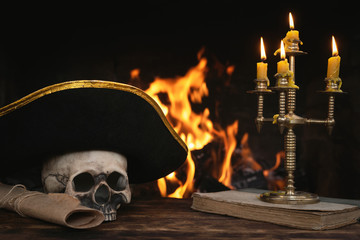 Wall Mural - Pirate table with human skull, captain hat, book and scroll map on a burning fire background. Piracy concept.