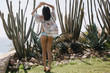 Full-length portrait from back of tanned brunette girl standing on tiptoe on cactus background. Slim stylish young woman with short dark hair looking at green peyote growing near the sea