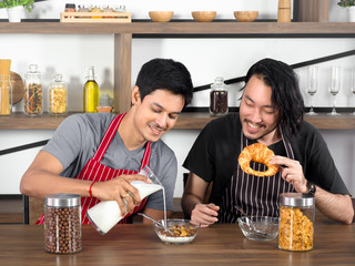 Pretty young lover having breakfast,one man pouring milk into cereal and the other holding croissants. Asian gay couple spending time together at home, LGBTQ+