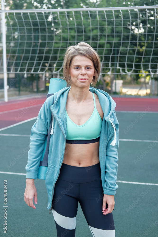 A woman in sports clothes and a yoga mat looks straight at the camera. A woman after a morning workout on the playground. A woman holding a gym mat and practicing yoga.
