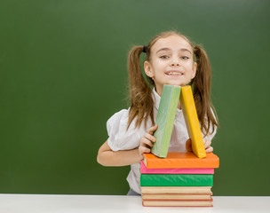 Happy little girl with books near empty green chalkboard. Empty space for text