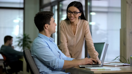 Wall Mural - Smiling business lady looking at male colleague sitting laptop, communication