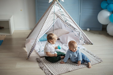 Two little infant male kids playing in toy tent indoor, in a pastel coloured playing in room, learning to smile, roll over, sit up, wave, clap, pick objects up.