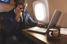 Young Attractive And Successful African American Businessman With Glassies Talking On The Phone And Working While Sitting In The Chair Of His Private Business Plane