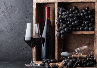 Bottle of red wine with dark grapes inside vintage wooden box on black stone background. Elegant wine glass with corks and corkscrew on black board.Natural light